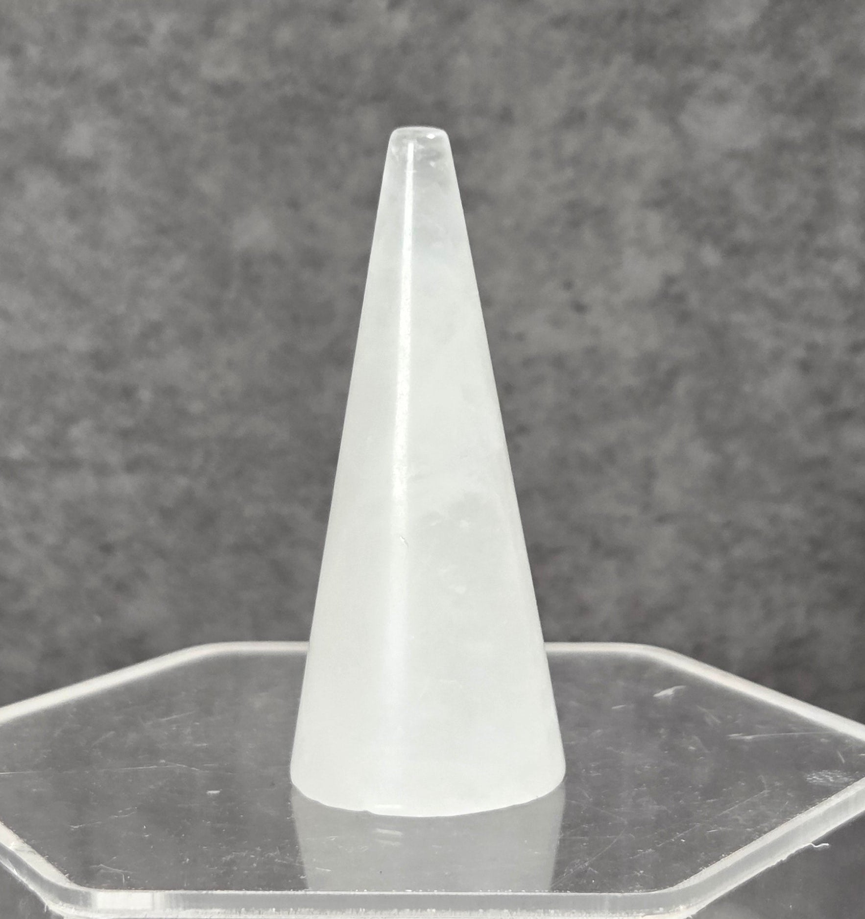 A frosted white cone-shaped object displayed upright on a clear hexagonal acrylic stand against a textured gray background.