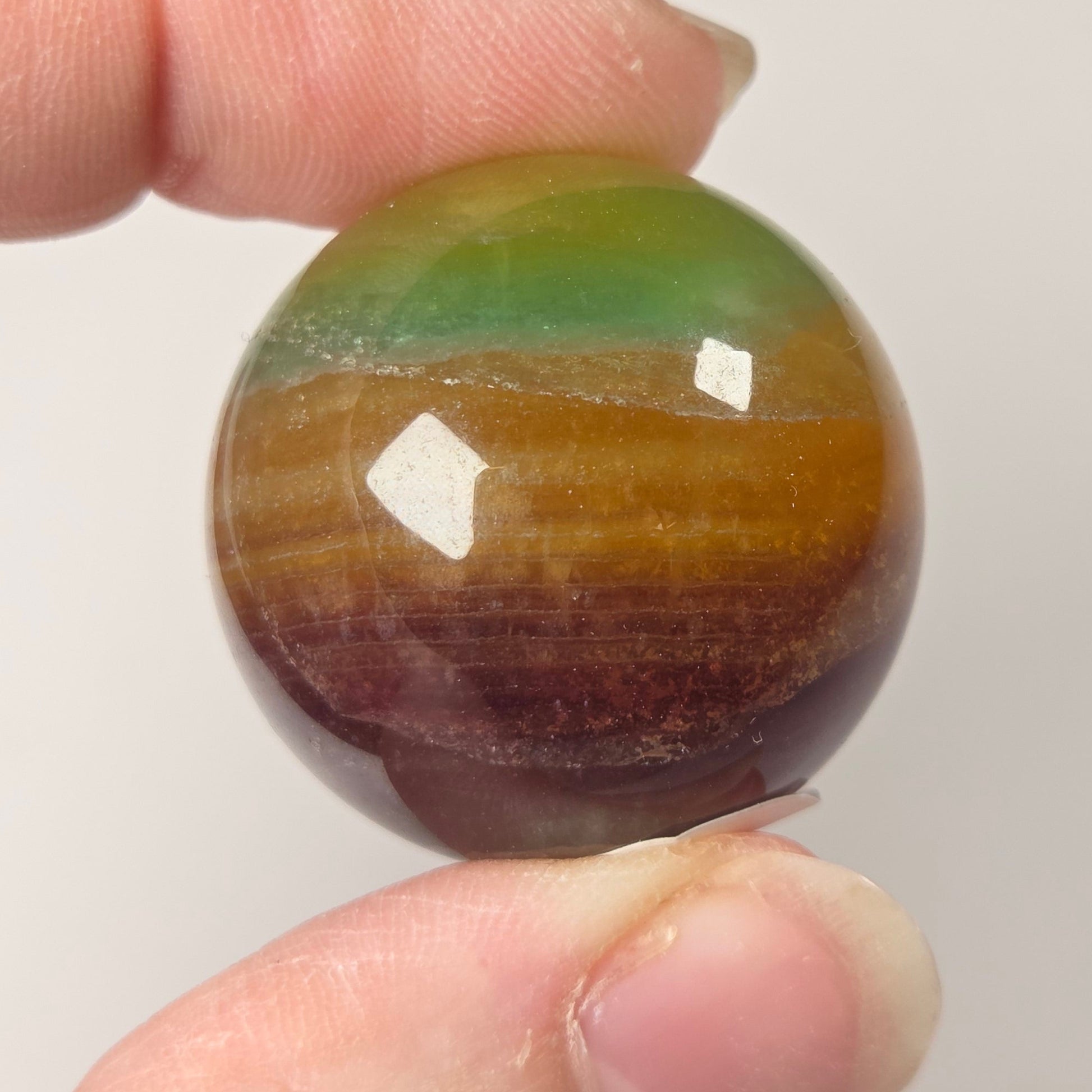 A close-up of a hand holding a polished round stone bead with a striped pattern of green, brown, and amber tones against a plain light background.