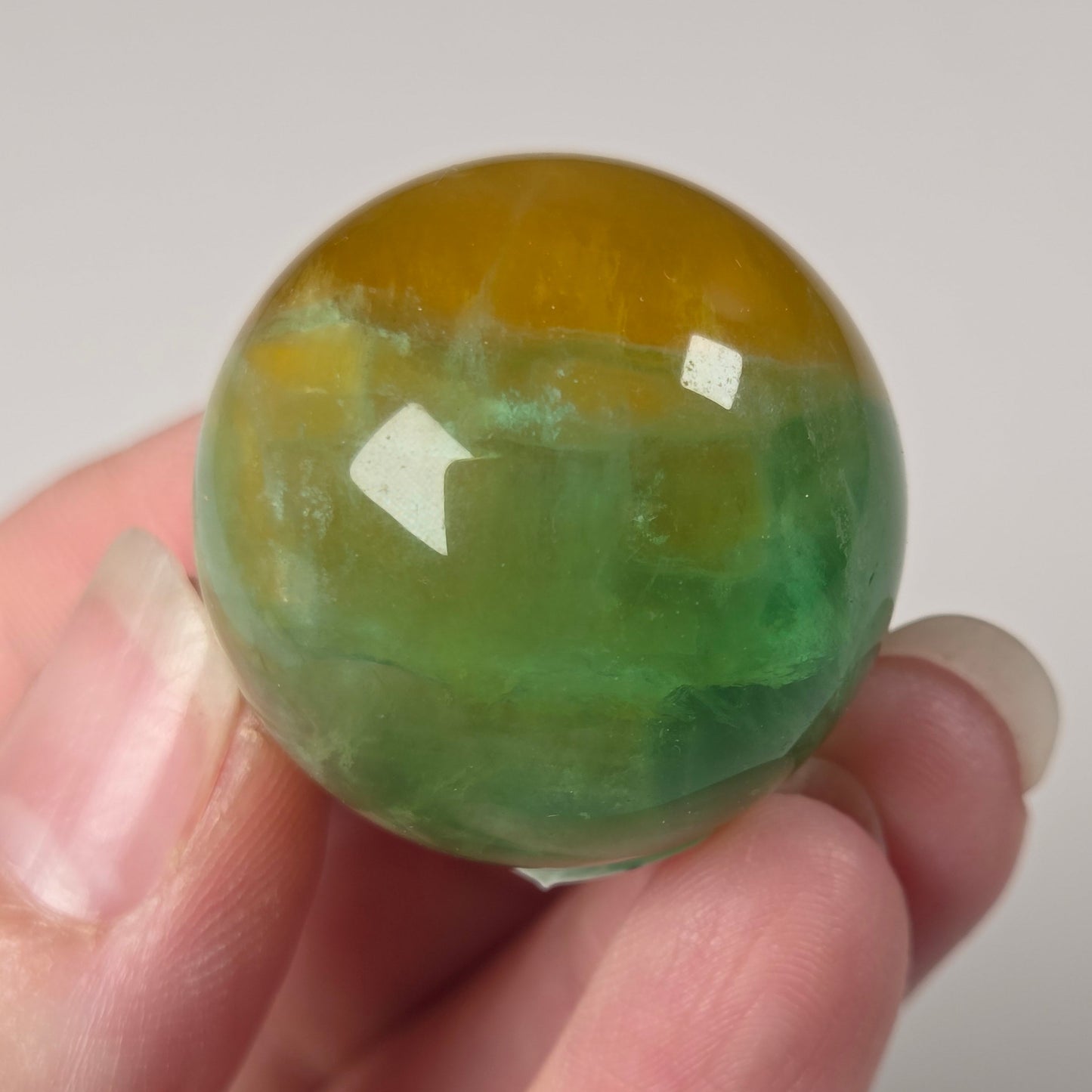 A hand holding a polished round stone sphere with a marbled green and yellow-orange pattern, set against a plain light gray background.