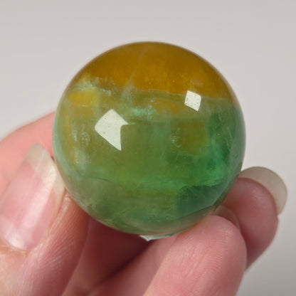 A hand holding a polished round stone sphere with a marbled green and yellow-orange pattern, set against a plain light gray background.