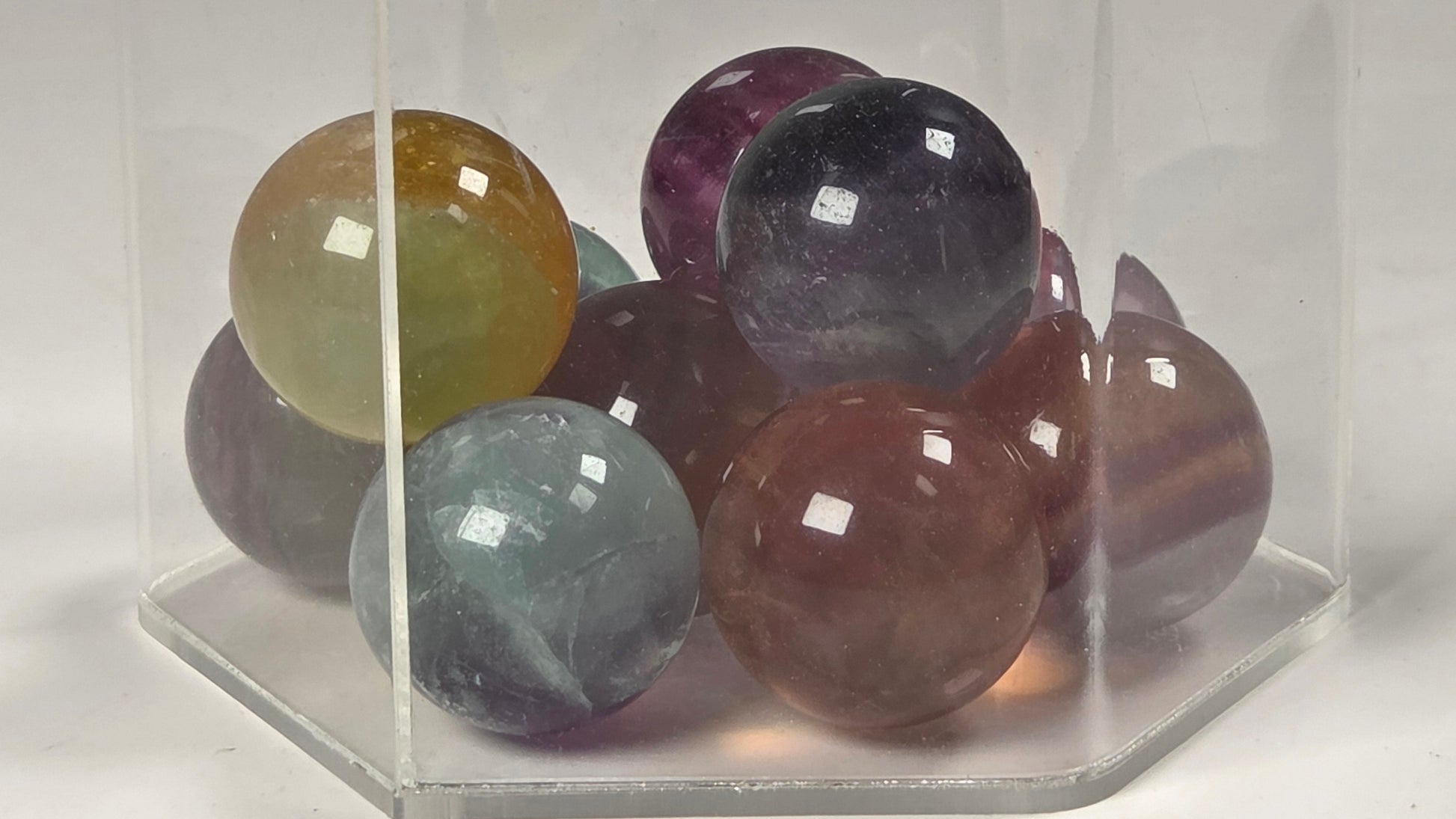 A group of polished multicolored gemstone spheres arranged inside a clear acrylic hexagonal display box on a white background.