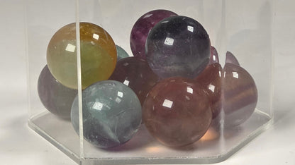 A group of polished multicolored gemstone spheres arranged inside a clear acrylic hexagonal display box on a white background.