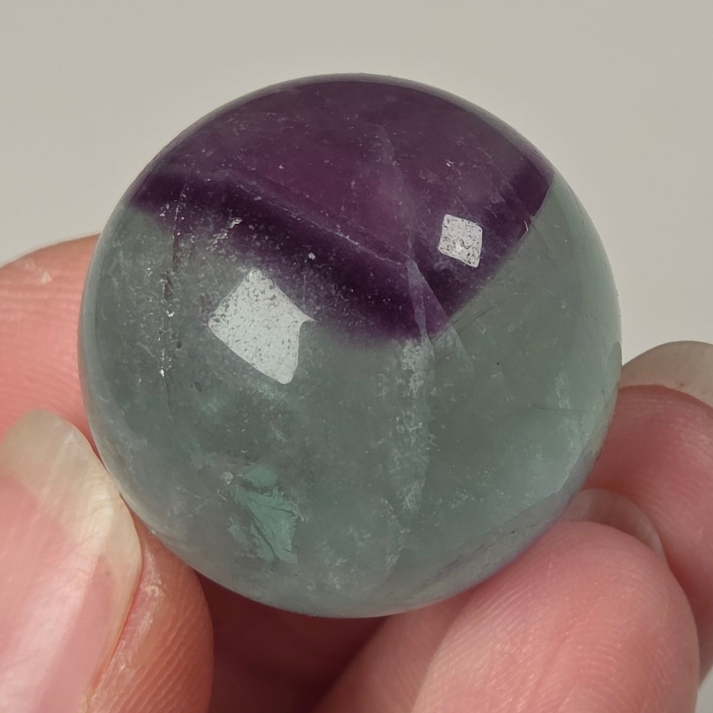 A hand holding a polished round gemstone with a two-tone color pattern of purple and pale gray-green, resembling a fluorite orb.
