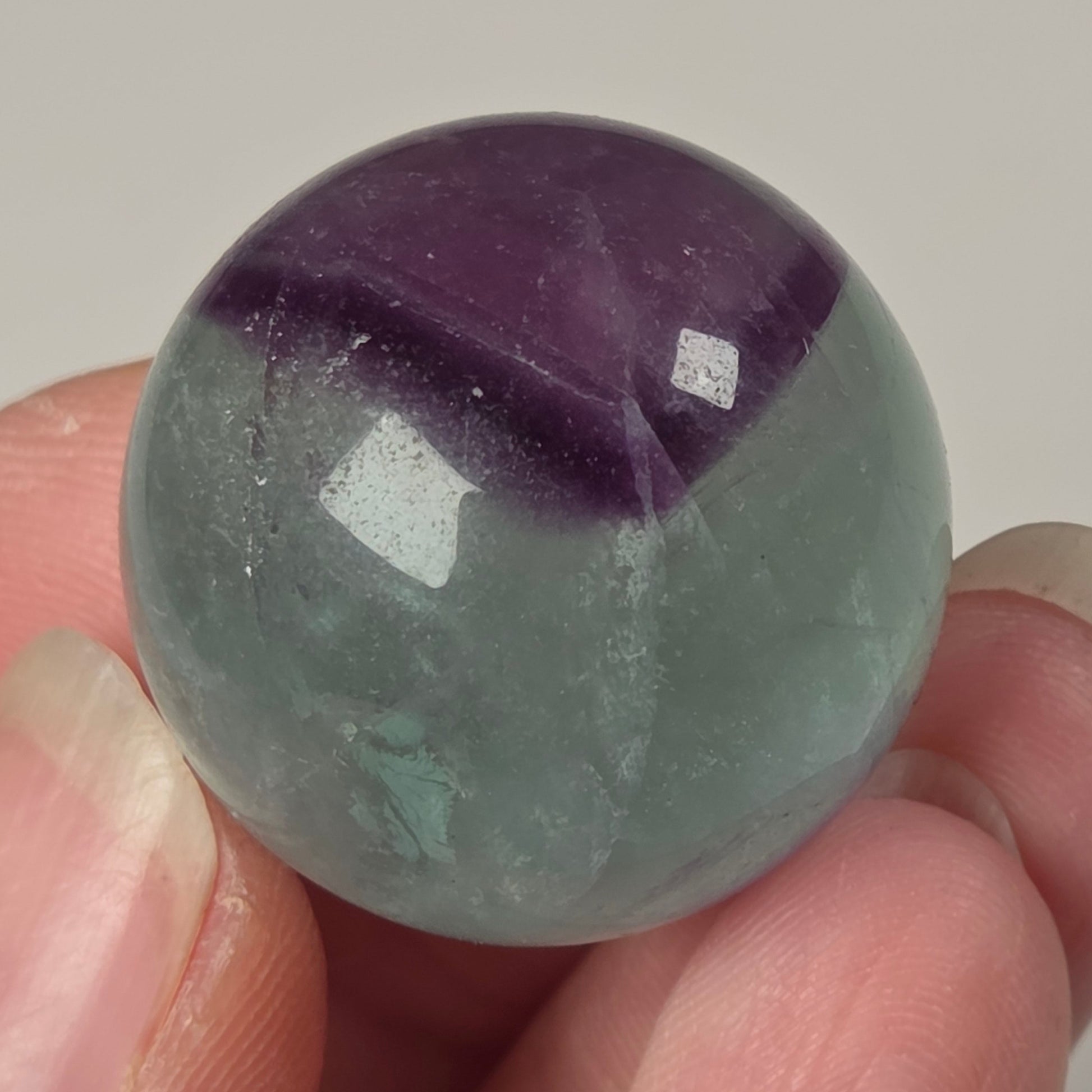 A hand holding a polished round gemstone with a two-tone color pattern of purple and pale gray-green, resembling a fluorite orb.