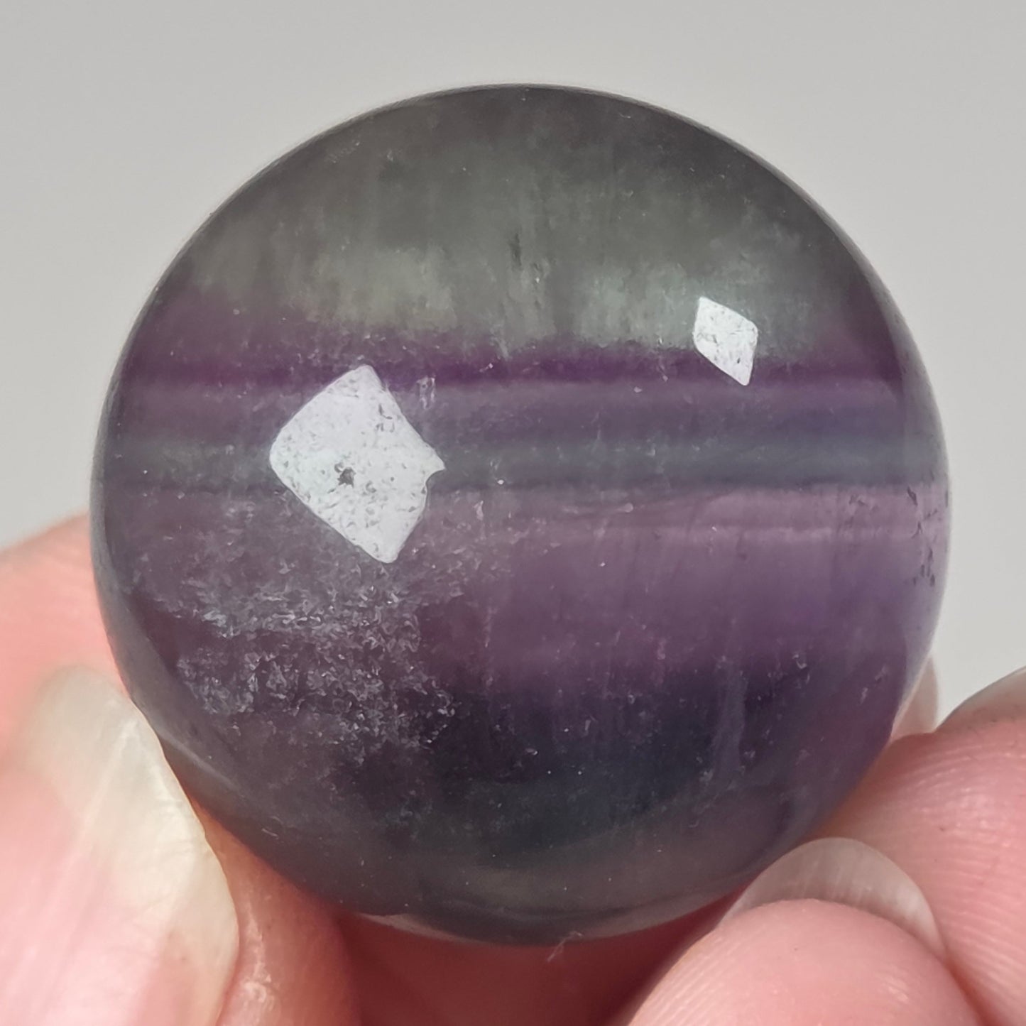 A polished purple and gray stone sphere held between fingers against a plain light background.