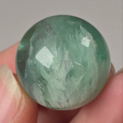 A close-up of a hand holding a polished round green stone bead or cabochon with visible internal crystalline structure against a plain light gray background.