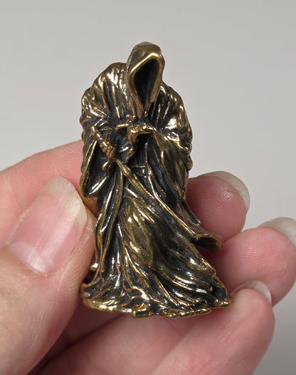 A hand holding a small, textured gold-toned metal charm or pendant with a leaf-like, organic shape against a plain light gray background.
