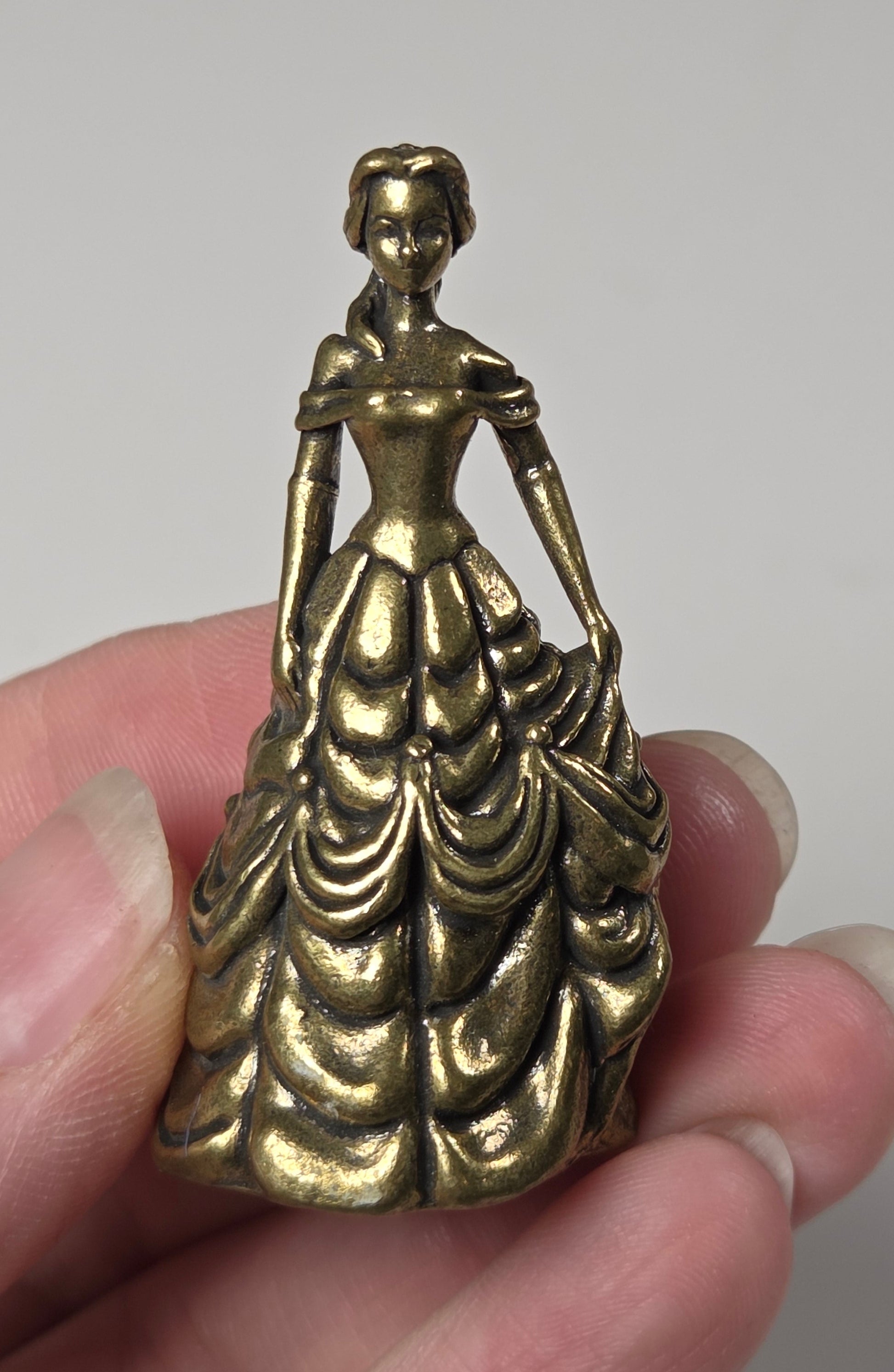 A small antique brass figurine of a classical female figure in a flowing dress, held in a person's hand against a plain light gray background.