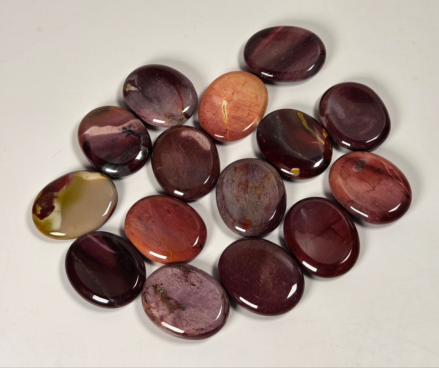 A group of polished oval cabochon gemstones in shades of deep burgundy, plum, reddish-brown, and one tan stone, arranged on a plain off-white background.