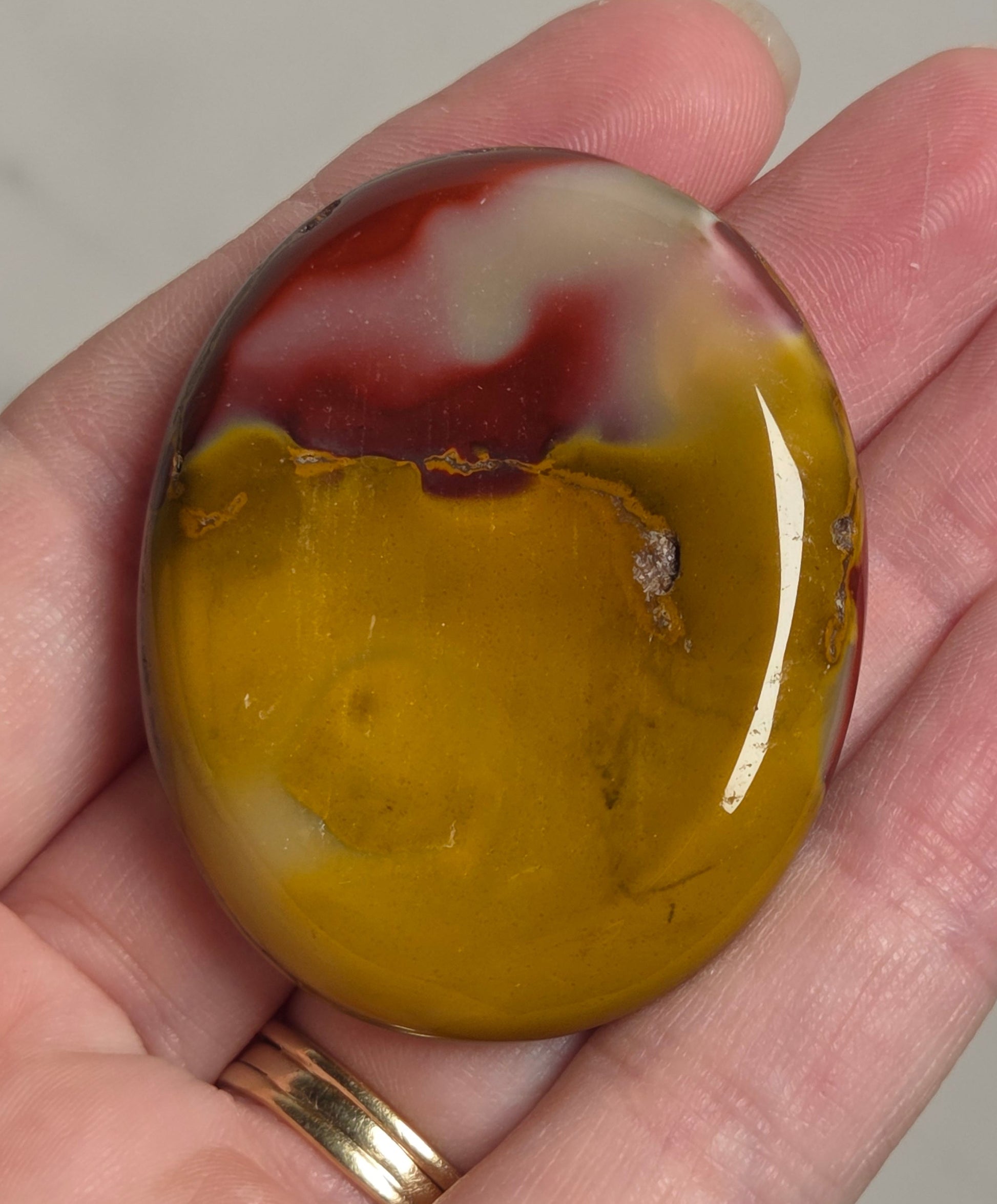 A polished oval cabochon gemstone with a marbled pattern of yellow, white, and red, held in a person's hand.