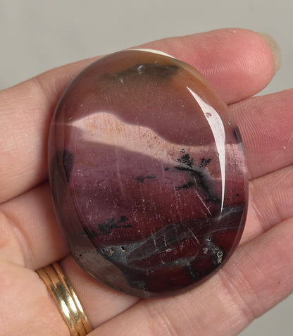 A polished oval tumbled stone with a deep reddish-brown and purple marbled pattern, held in a person's hand.