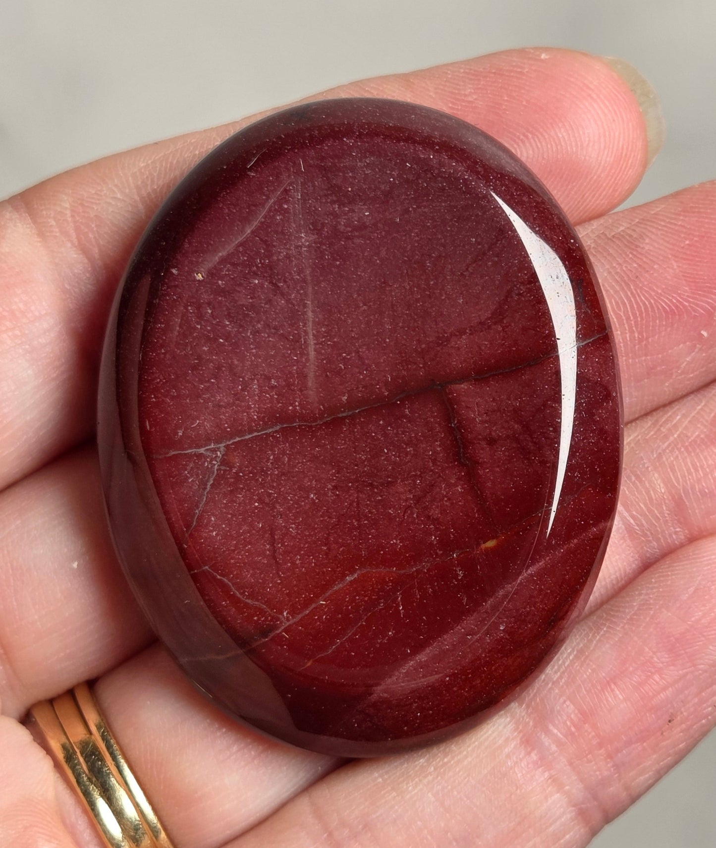 A polished oval red stone or cabochon held in a person's hand, with a gold ring visible on the wrist against a light gray background.