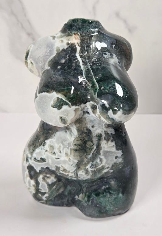 A polished natural stone figurine of a woman's torso, showcasing a mix of dark forest green, glossy black, and white marbled patterns, set against a split white and light gray marble background.