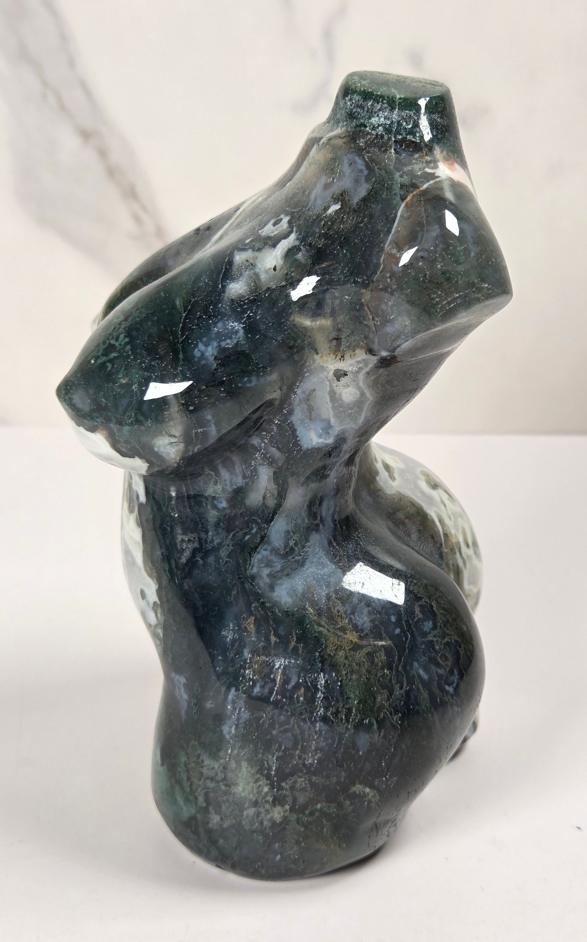 A polished natural stone sculpture of a stylized female torso, made from dark green and gray marbled stone with white veining, shown against a white marble background.