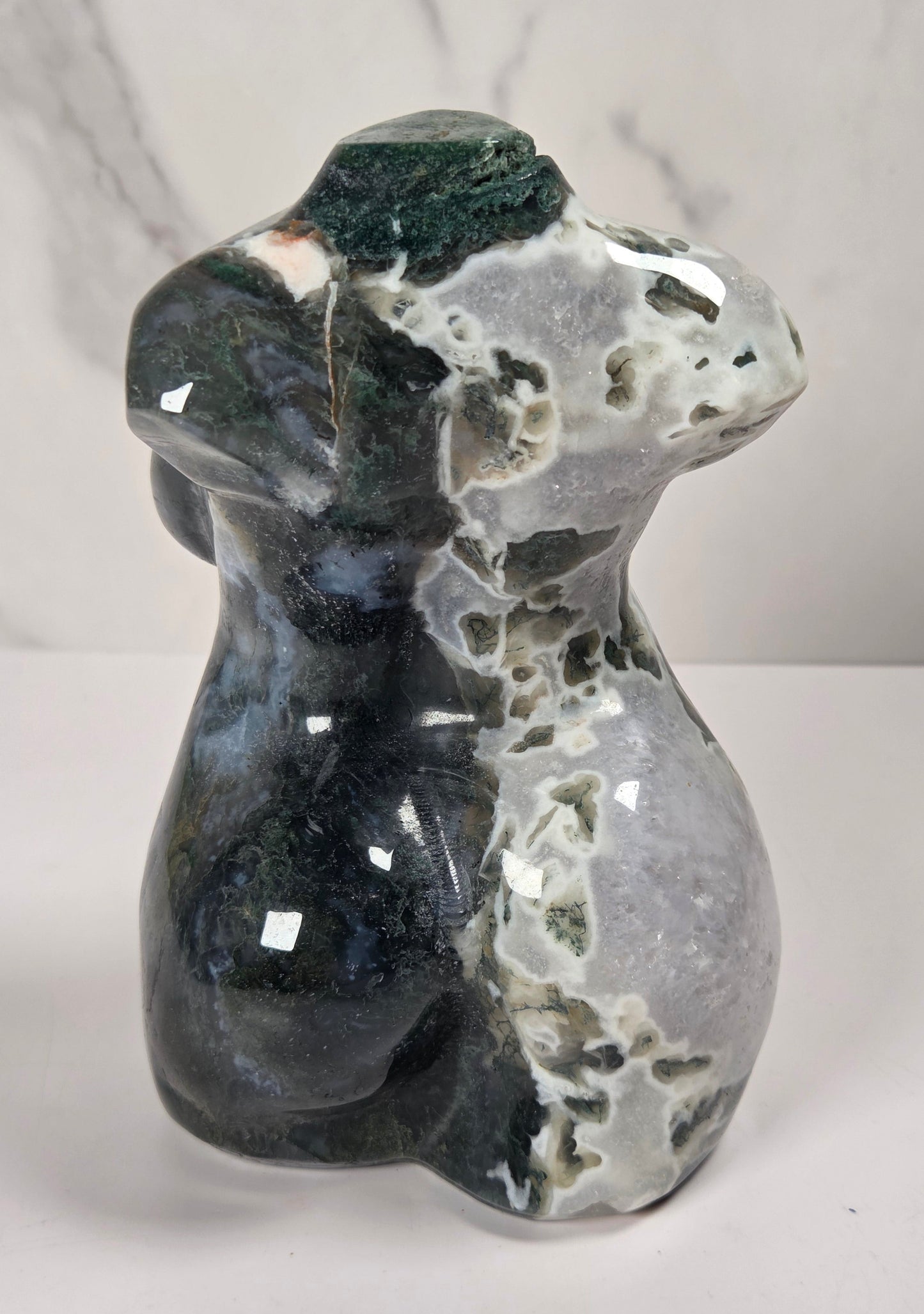A polished natural stone figurine shaped like a female torse, featuring a mottled pattern of black, gray, white, and dark green, displayed on a light marble-patterned surface.