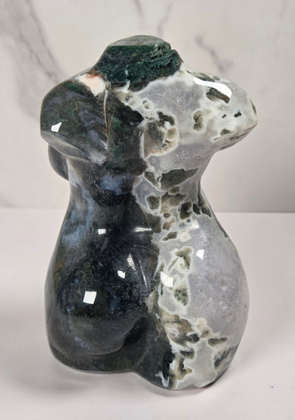 A polished natural stone figurine shaped like a female torse, featuring a mottled pattern of black, gray, white, and dark green, displayed on a light marble-patterned surface.
