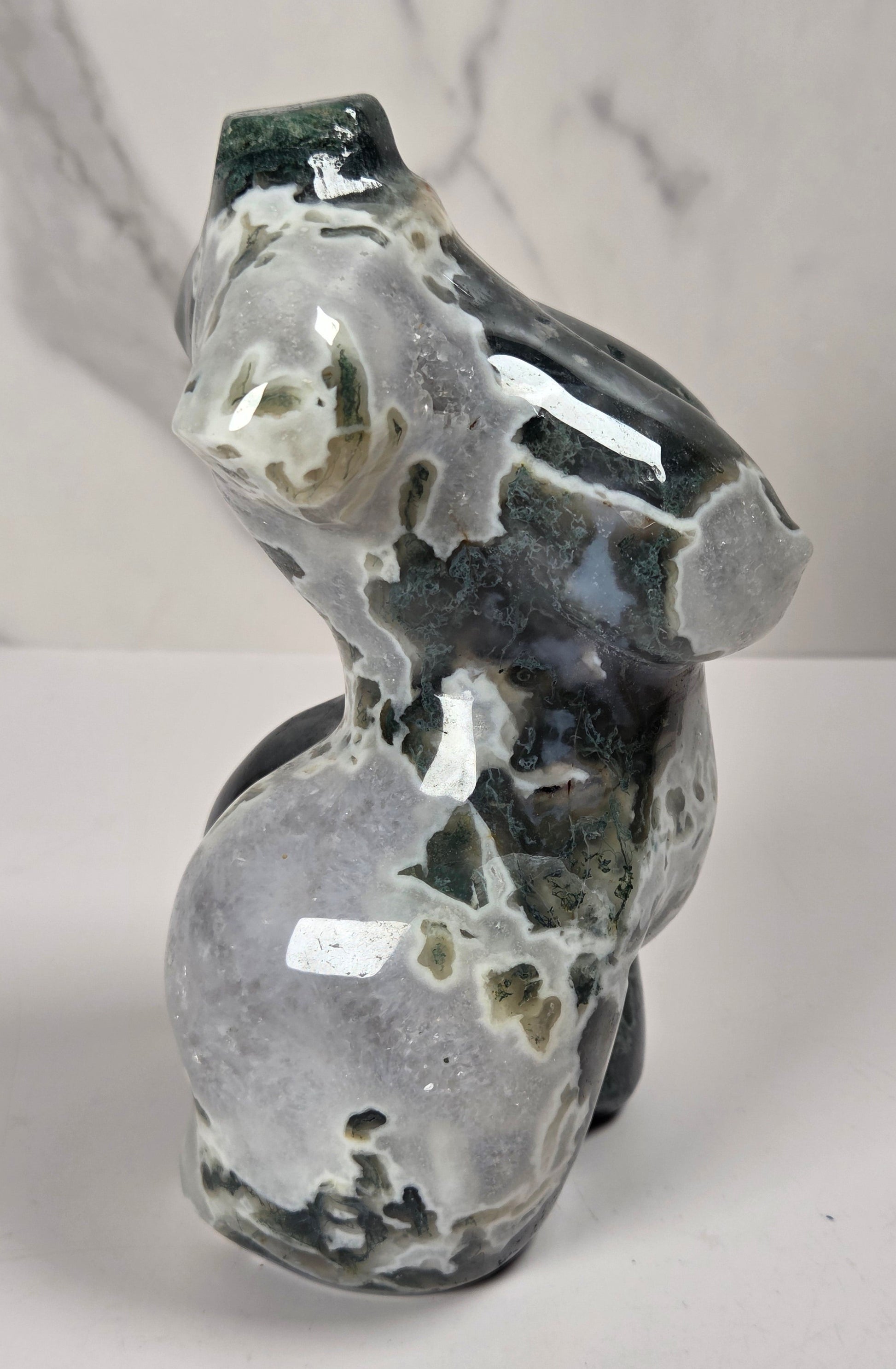 A polished natural stone sculpture shaped like a stylized female torso, shown against a white marble-patterned background.