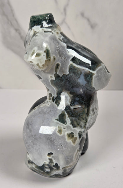 A polished natural stone sculpture shaped like a stylized female torso, shown against a white marble-patterned background.