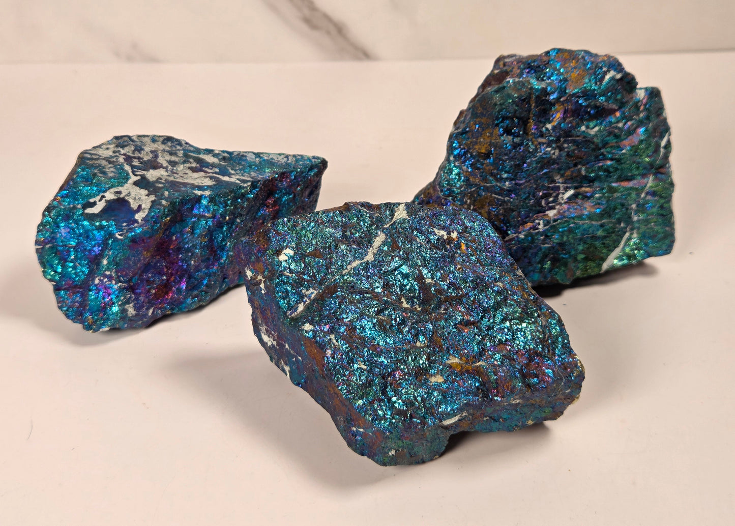 Three rough, iridescent blue and teal mineral specimens with metallic shimmer, displayed on a light beige surface against a white marble-patterned background.