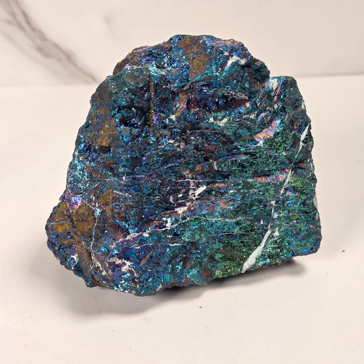 A raw, rough mineral specimen with a highly iridescent, metallic surface showing shimmering blue, green, and purple hues, set against a plain off-white background.