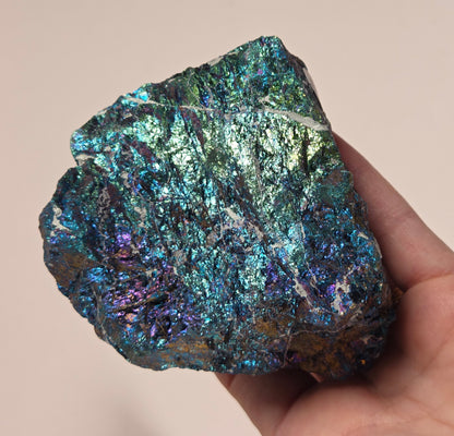A hand holding an iridescent mineral specimen with shimmering blue, green, and purple tones against a plain light beige background.