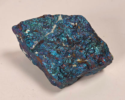 A raw, rough mineral specimen with a highly textured, metallic surface showing vibrant blue, teal, and dark brown iridescent hues, set against a plain light beige background.