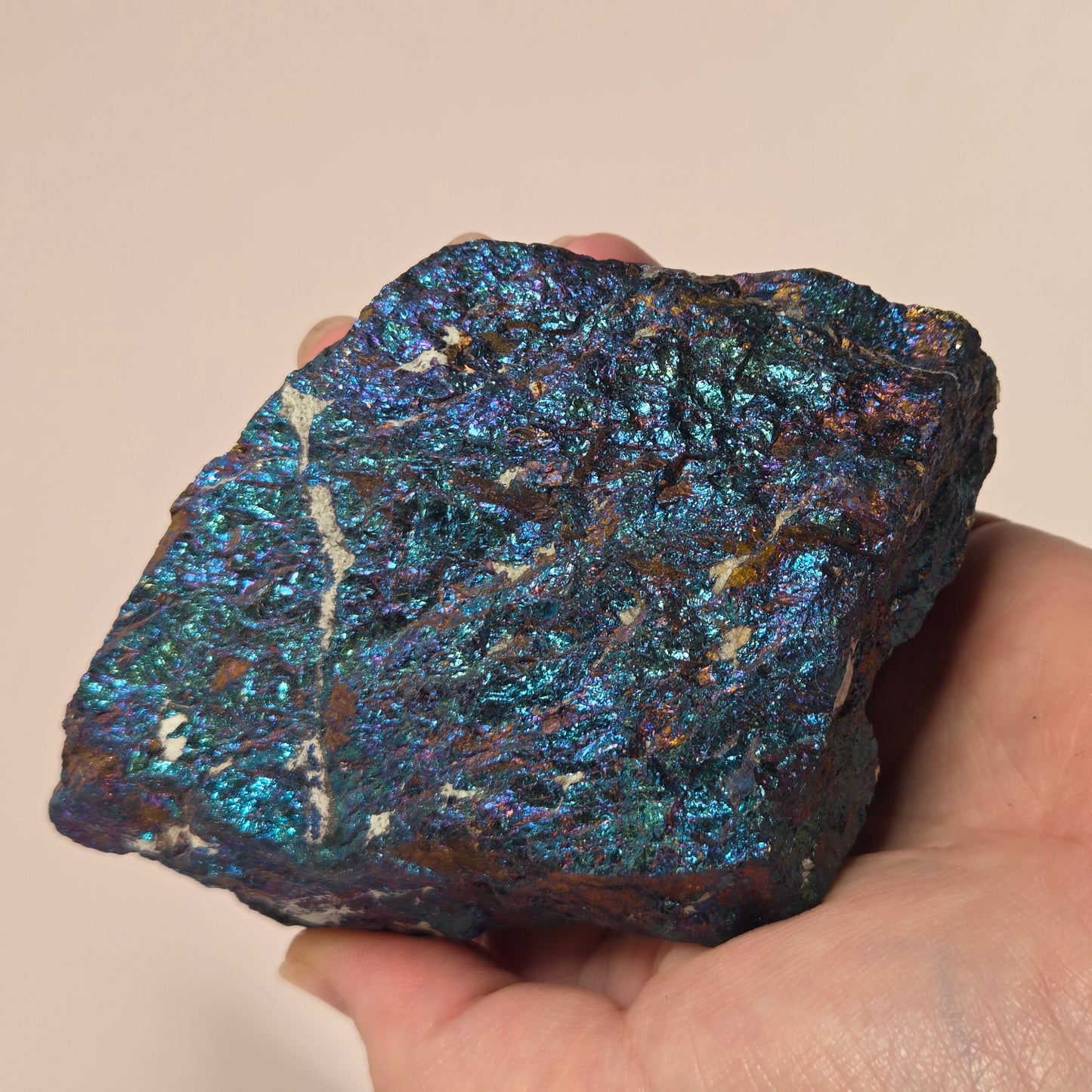 A hand holding an iridescent blue and green metallic mineral specimen against a plain beige background.