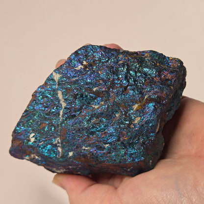 A hand holding an iridescent blue and green metallic mineral specimen against a plain beige background.