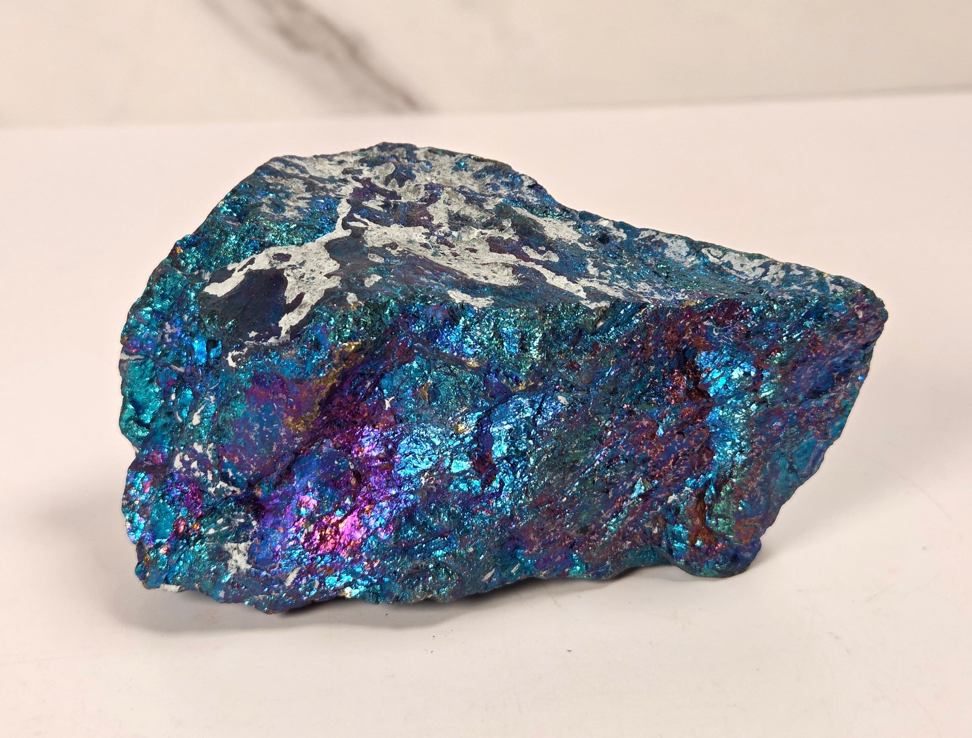 A raw, rough mineral specimen with a metallic, iridescent sheen displaying blue, teal, purple, and silver colors, set against a light gray marble-patterned background.