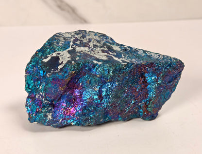 A raw, rough mineral specimen with a metallic, iridescent sheen displaying blue, teal, purple, and silver colors, set against a light gray marble-patterned background.
