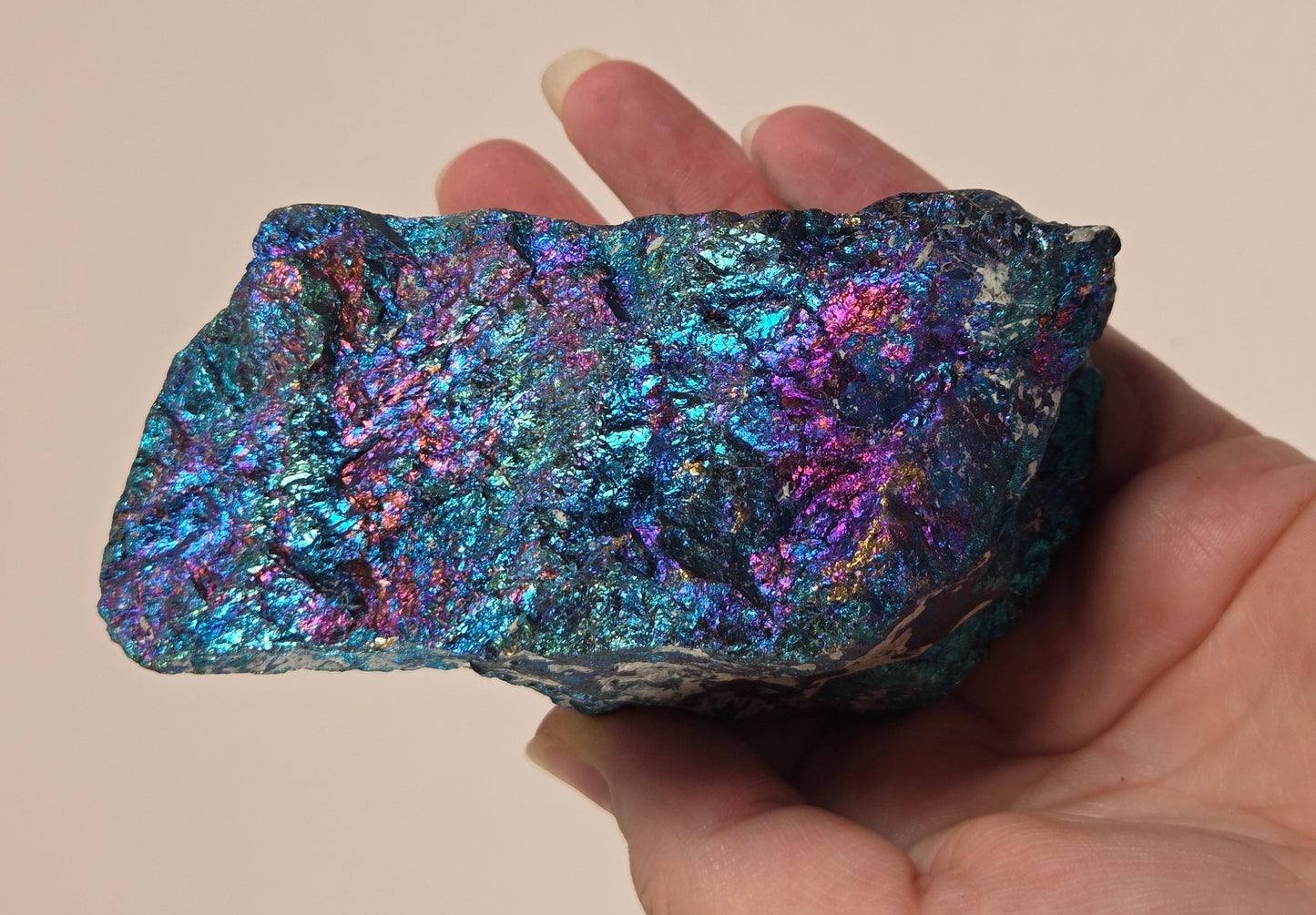 A hand holding an iridescent metallic mineral specimen showing shimmering blue, purple, and pink tones against a plain beige background.