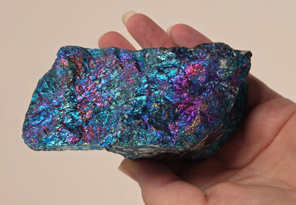 A hand holding an iridescent metallic mineral specimen showing shimmering blue, purple, and pink tones against a plain beige background.