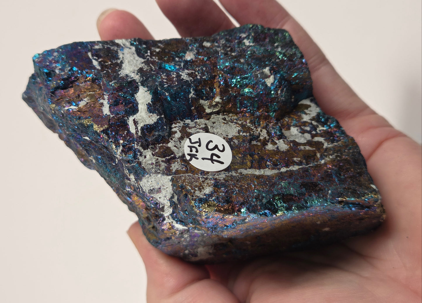 A hand holding an iridescent mineral specimen with a small white sticker labeled '34 254' on its surface, set against a plain light background.