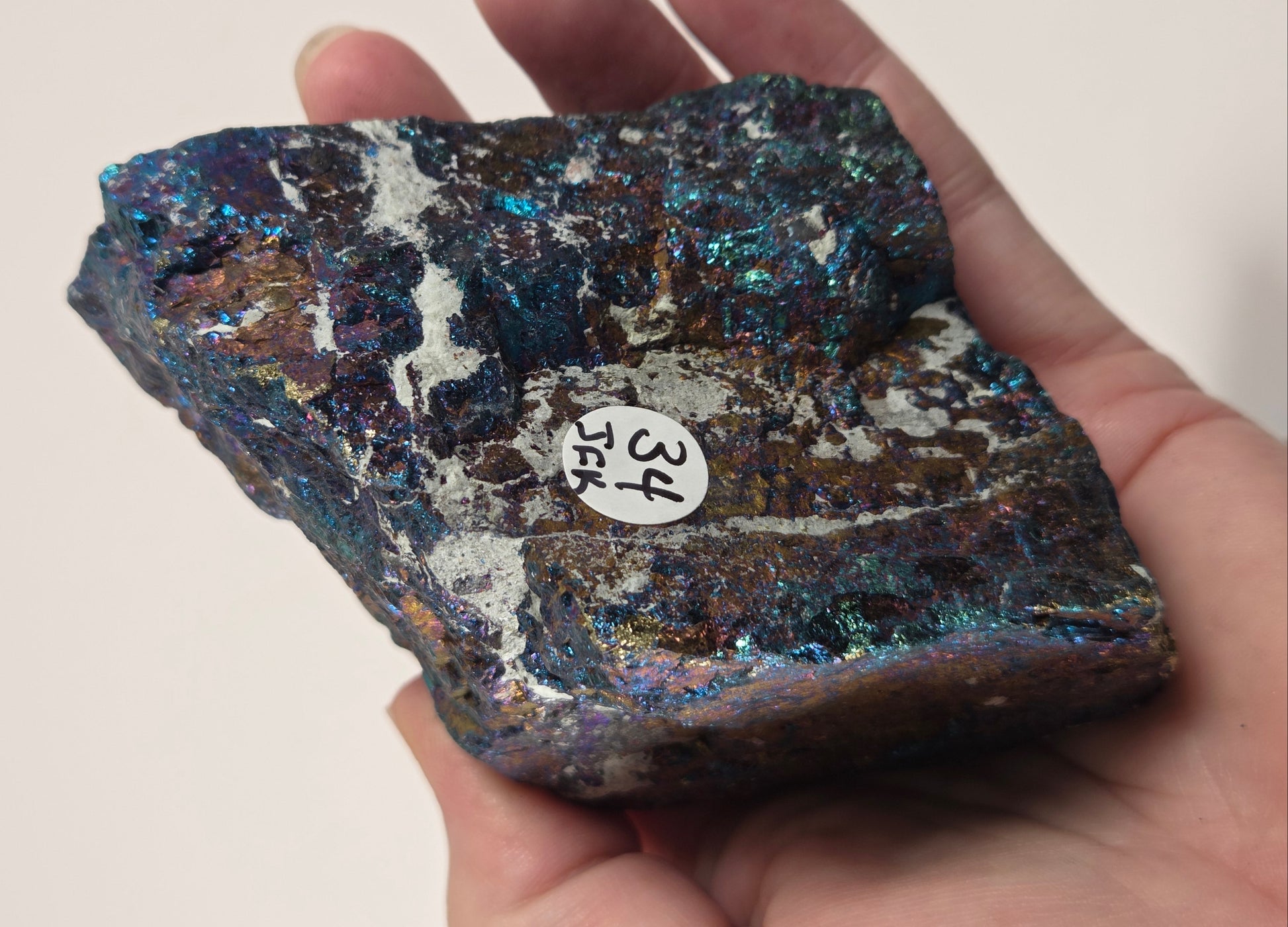 A hand holding an iridescent mineral specimen with a small white sticker labeled '34 254' on its surface, set against a plain light background.