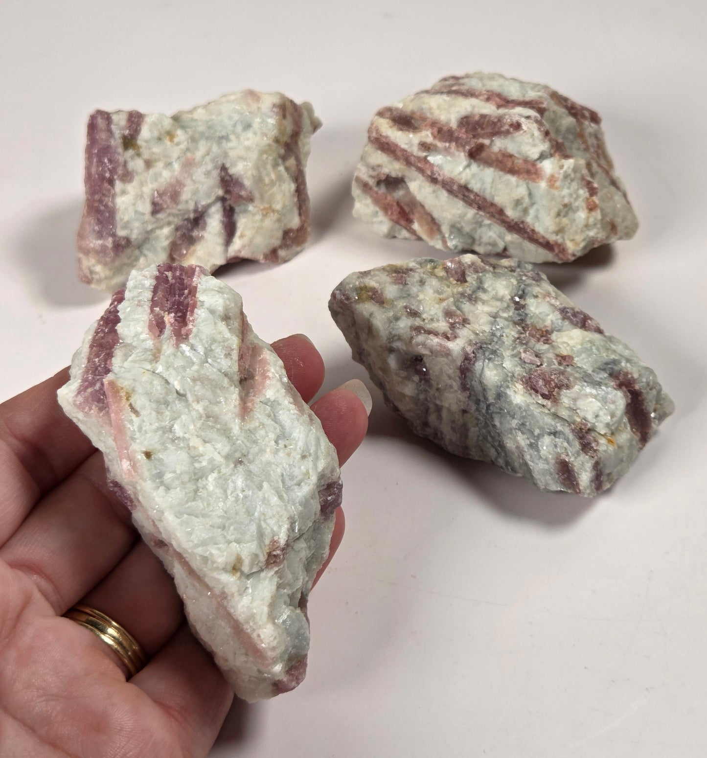 Four rough mineral or stone pieces with pale greenish-gray and dusty rose/brown veining, with one held in a person's hand against a plain light background.