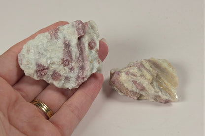 A hand holding a rough, pale pink and white mineral or rock specimen, with a second similar specimen hanging on a plain light wall above.