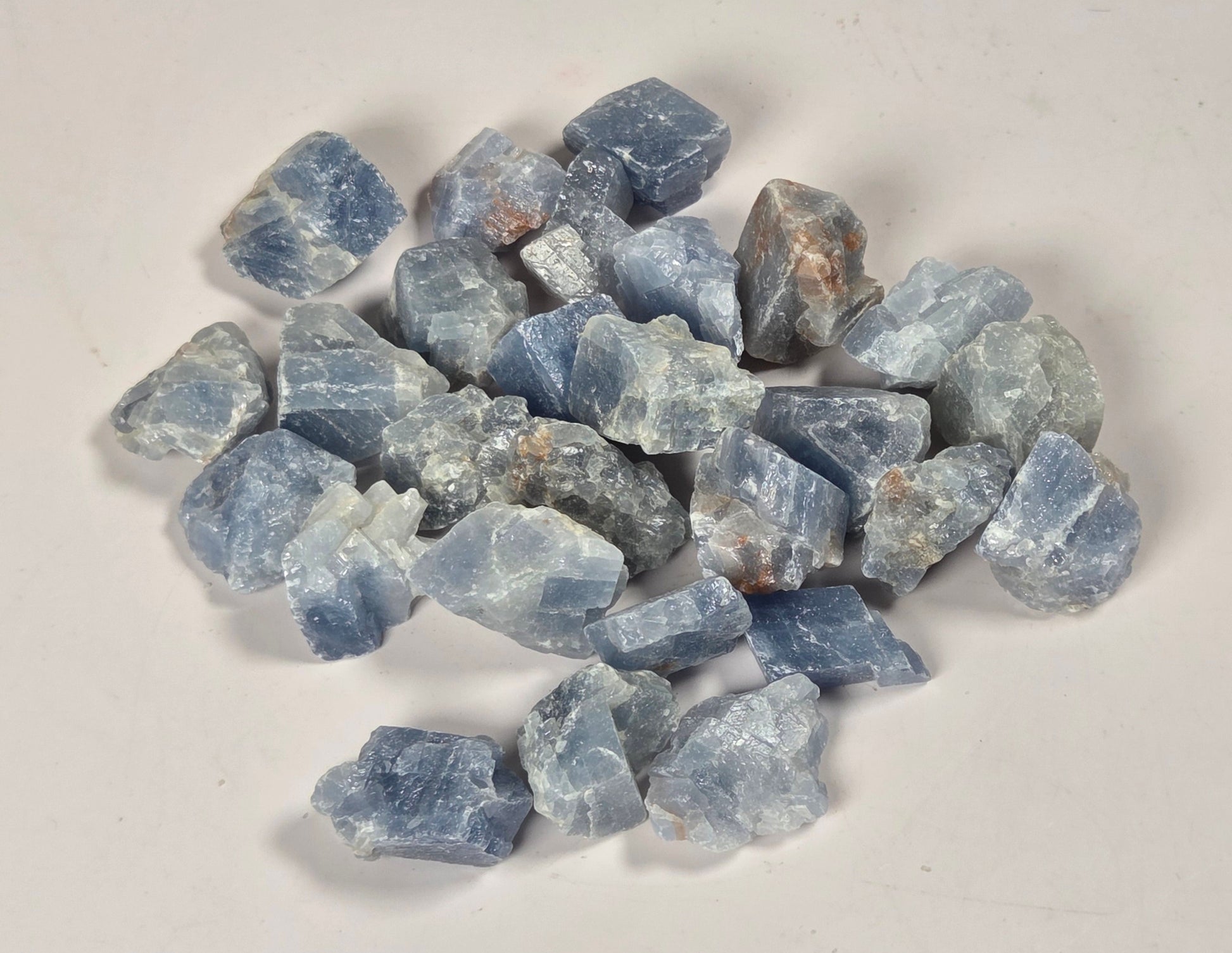 A pile of rough, unpolished blue gemstone chips with natural crystalline textures, arranged on a plain off-white surface.