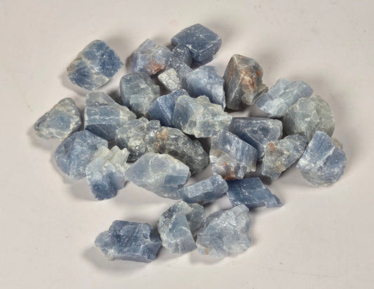 A pile of rough, unpolished blue gemstone chips with natural crystalline textures, arranged on a plain off-white surface.