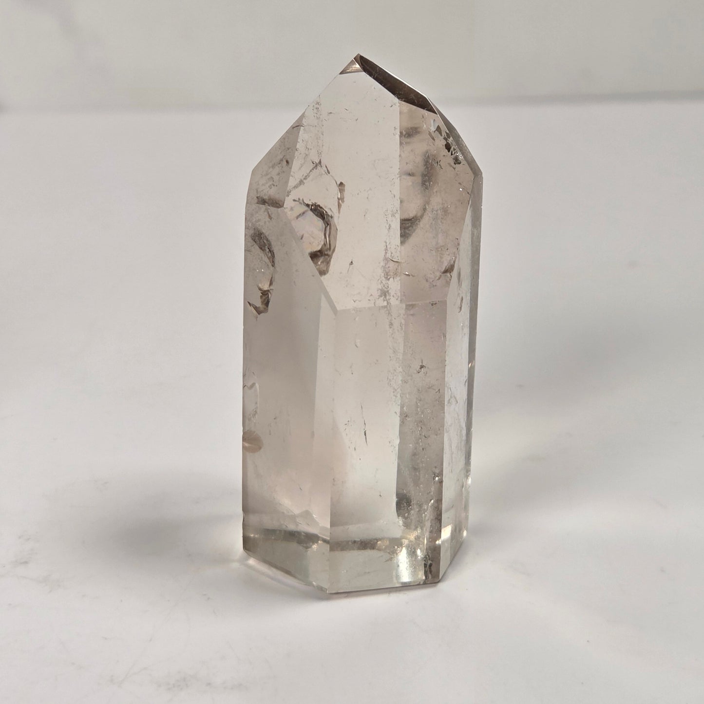 A clear quartz crystal point with natural inclusions, standing upright on a white surface against a light gray background.