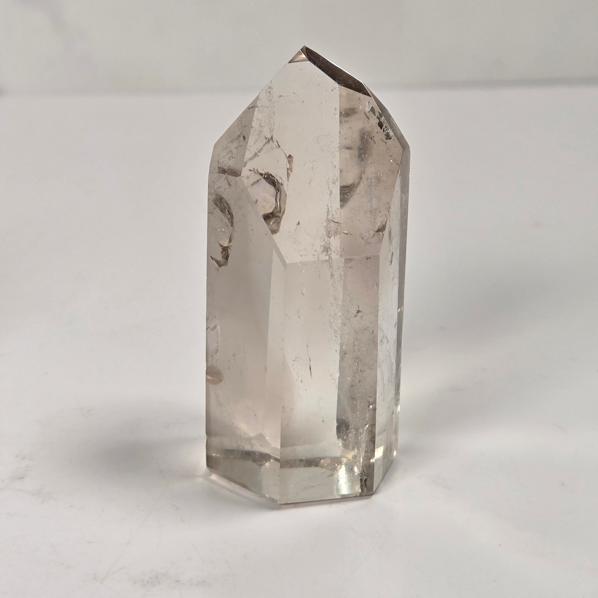 A clear quartz crystal point with natural inclusions, standing upright on a white surface against a light gray background.