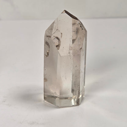 A clear quartz crystal point with natural inclusions, standing upright on a white surface against a light gray background.
