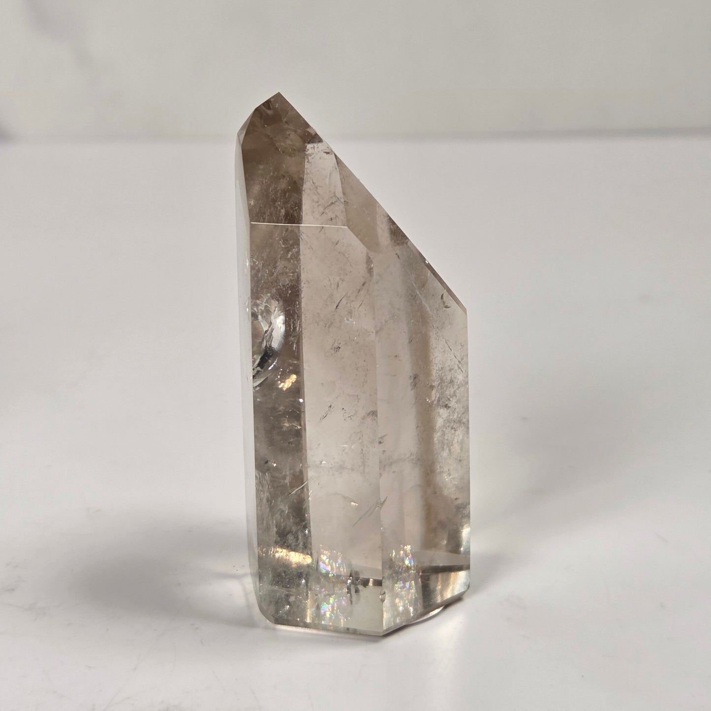 A natural clear quartz crystal point with internal inclusions, standing upright on a light gray surface against a light gray background.