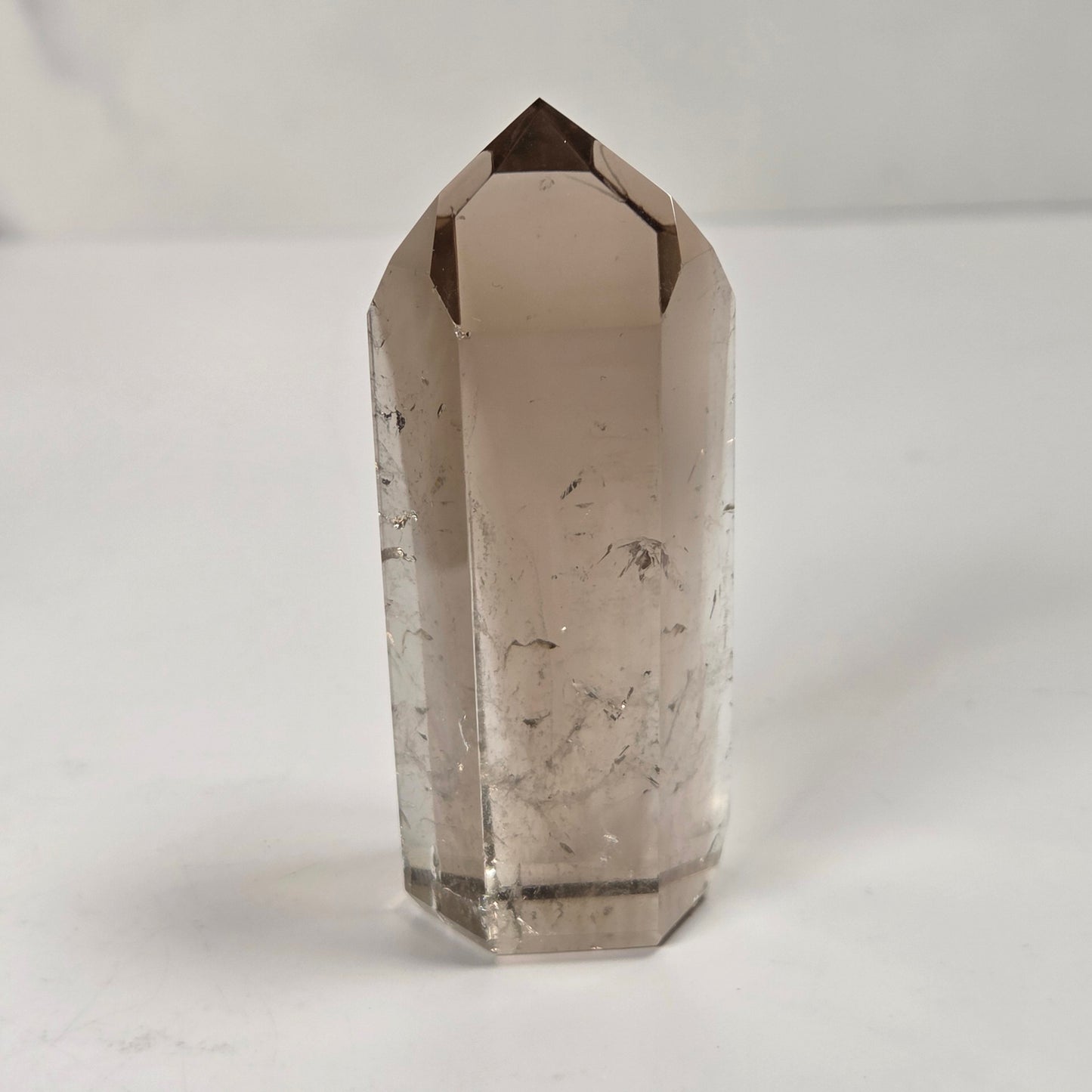 A natural smoky quartz crystal point with visible inclusions, standing upright on a white surface against a light gray background.