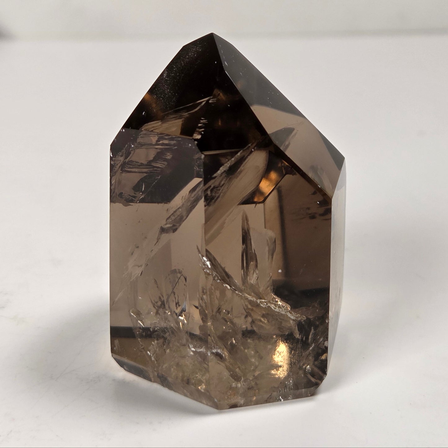 A natural smoky quartz crystal point with a hexagonal shape, showing translucent brown and gray tones, photographed on a light gray surface.