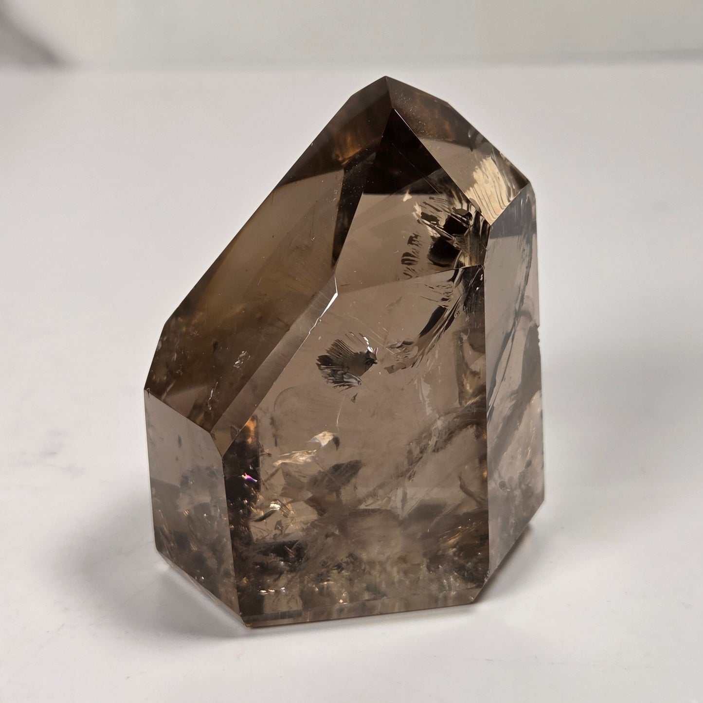 A natural smoky quartz crystal point with a hexagonal shape, showing internal inclusions and a translucent brown-gray color, photographed on a plain light gray surface.