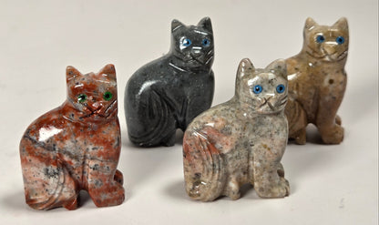 Four small carved cat figurines made from polished stone, each with different natural stone patterns and blue glass eyes, arranged on a light gray background.