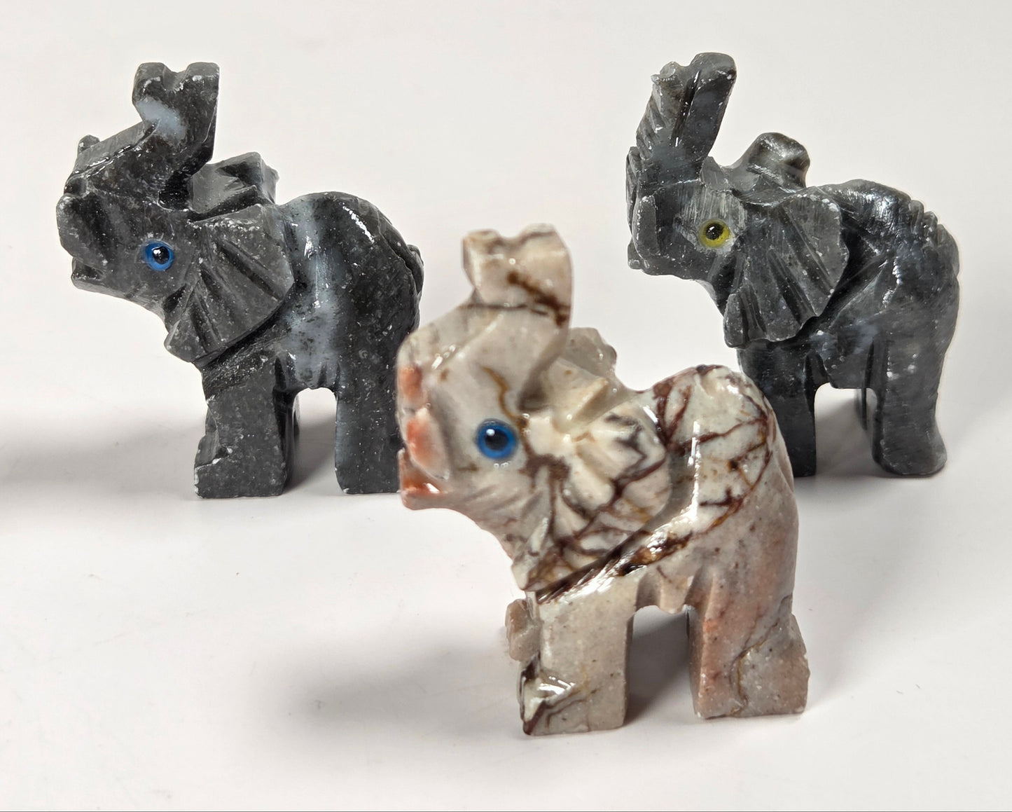 Three small elephant figurines made from natural stone, including two dark gray elephant statues and one light beige elephant with brown veining and blue eyes, displayed on a white background.