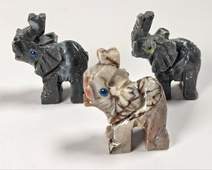 Three small elephant figurines made from natural stone, including two dark gray elephant statues and one light beige elephant with brown veining and blue eyes, displayed on a white background.