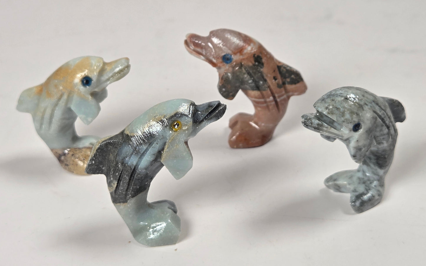 Four small carved dolphin figurines in different colors and materials, including light blue, brown, gray, and beige, arranged on a plain light background.