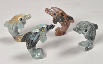 Four small carved dolphin figurines in different colors and materials, including light blue, brown, gray, and beige, arranged on a plain light background.